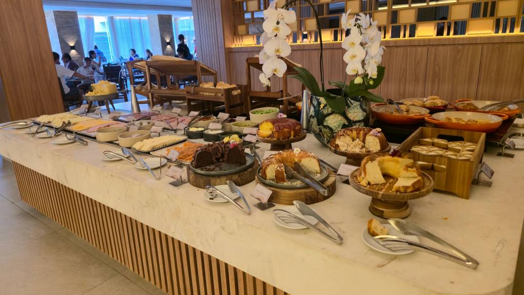 Um buffet de café da manhã com diversas opções, incluindo bolos, pães, frios, frutas e pratos quentes, em um ambiente de restaurante com pessoas ao fundo