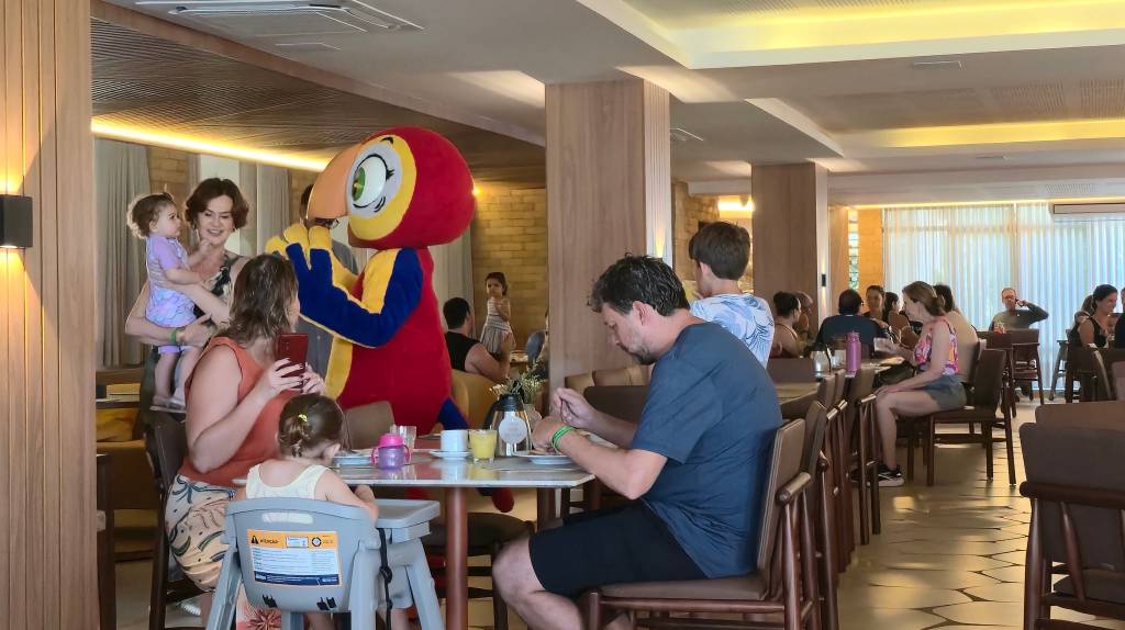 Pessoas em um restaurante com um mascote de papagaio vermelho e amarelo. Uma mulher segura um bebê, outra tira foto com celular, e um homem come à mesa.