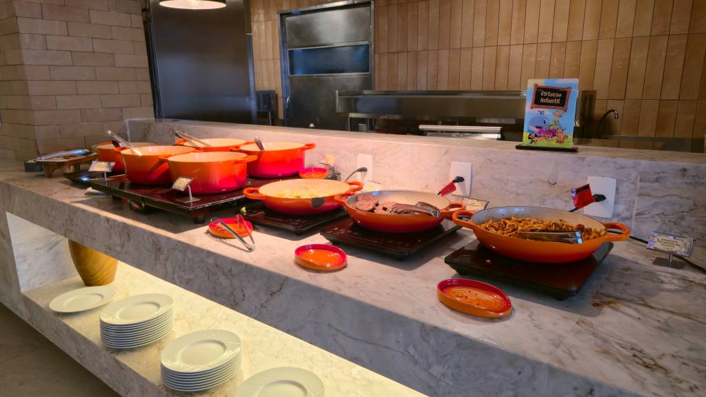 Um buffet com diversas panelas de ferro fundido laranja e vermelhas cheias de comida, sobre um balcão de mármore claro. Há pilhas de pratos brancos embaixo e um cartaz colorido de Estação Infantil ao fundo