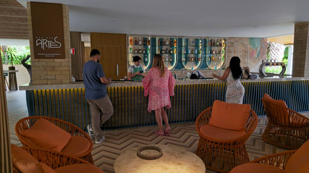 Pessoas em um bar moderno com decoração em tons terrosos e azuis. Um homem de camiseta azul e uma mulher de saída de praia rosa conversam com o bartender. Outra mulher de vestido branco estampado está no balcão. Poltronas de vime laranja com almofadas e uma mesa de centro de madeira completam o ambiente. Ao fundo, vegetação tropical.
