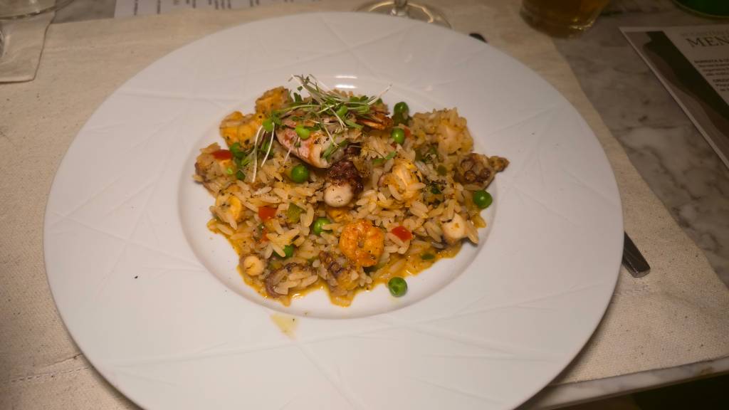 Arroz de frutos do mar com camarão, polvo, ervilhas e pimentão vermelho, decorado com brotos verdes, servido em um prato branco sobre uma toalha de mesa clara e um menu ao fundo