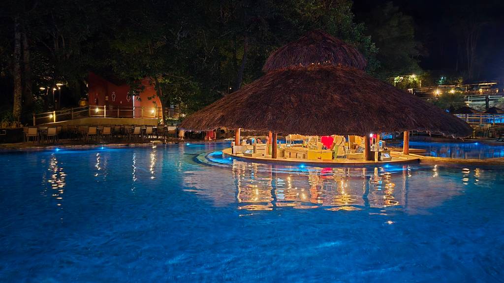 Piscina azul iluminada à noite com um bar central de telhado de palha, refletindo luzes na água. Ao fundo, uma construção avermelhada e árvores escuras, com mesas e cadeiras dispostas em um deck.