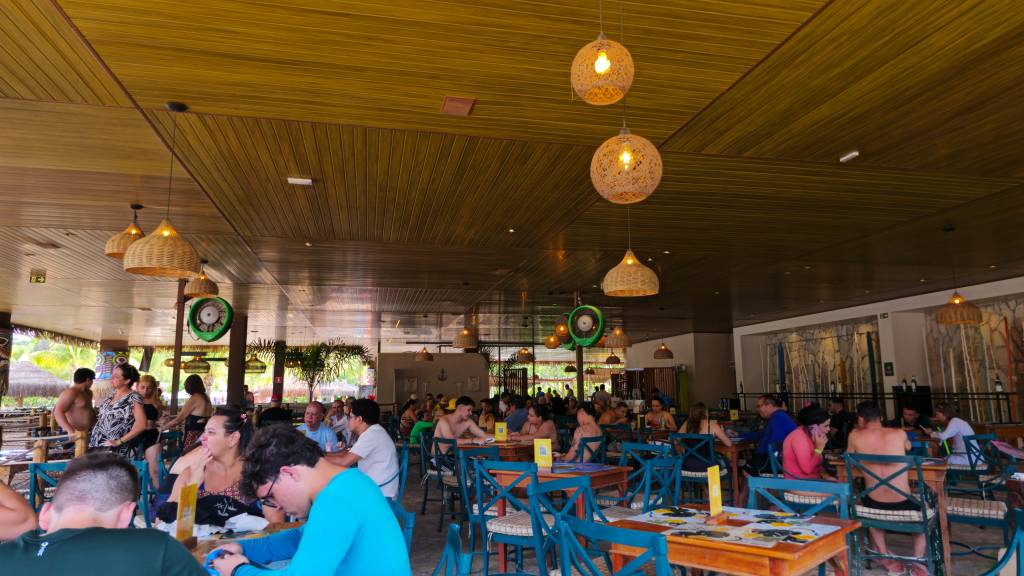 Pessoas sentadas em mesas de madeira com cadeiras azuis em um restaurante com teto de madeira e luminárias de palha, algumas conversando e outras comendo