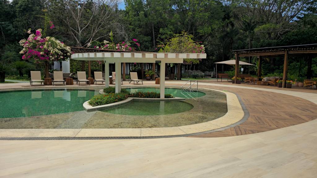 Piscina de resort com água verde-azulada, cercada por espreguiçadeiras sob um pergolado branco e árvores floridas. Ao fundo, um caminho de madeira e outro pergolado de madeira com bancos e mesas, em meio à vegetação densa