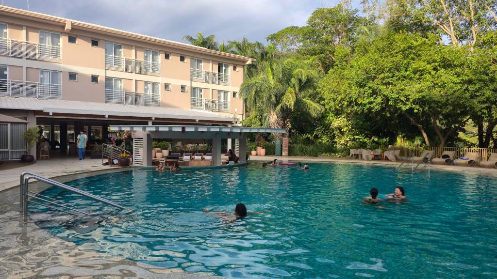 Piscina de um resort com pessoas nadando e relaxando. Ao fundo, um prédio de três andares com varandas, um bar molhado com bancos e espreguiçadeiras, e árvores exuberantes. O céu está parcialmente nublado, e a água da piscina é azul-turquesa