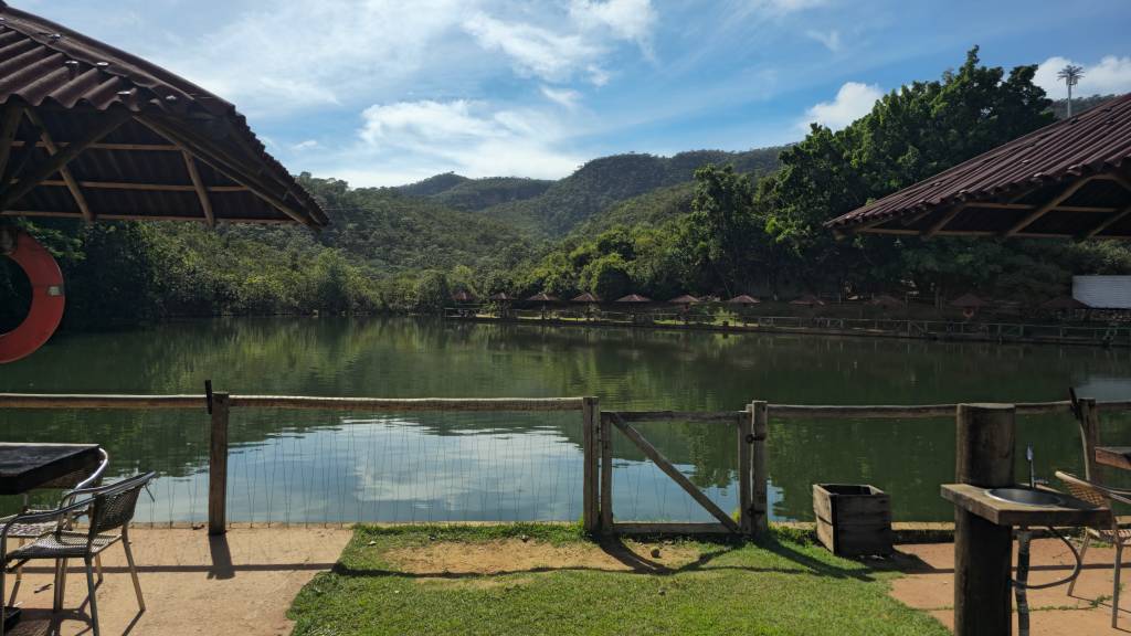 Vista de um lago verde-escuro com montanhas arborizadas ao fundo sob um céu azul com nuvens esparsas. Em primeiro plano, um cercado de madeira e tela, com um portão à direita. Há um quiosque com telhado de telhas de barro e um colete salva-vidas vermelho pendurado à esquerda. Na margem oposta, pequenos quiosques com guarda-sóis marrons sobre a água. À direita, uma pia rústica de madeira.