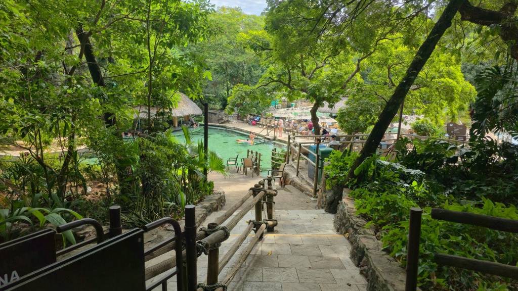 Escadaria de pedra com corrimão rústico de madeira, descendo para uma área de lazer com piscina de água verde-clara, rodeada por árvores frondosas e vegetação exuberante. Pessoas relaxam em espreguiçadeiras e dentro da piscina, sob guarda-sóis e quiosques de palha.
