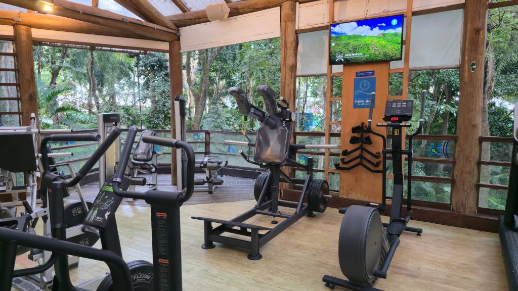 Academia ao ar livre com equipamentos de musculação e cardio, como elíptico e leg press, em um deck de madeira cercado por árvores e vegetação exuberante. Um monitor exibe uma paisagem verde com céu azul, e um aviso azul indica Tempo Livre.