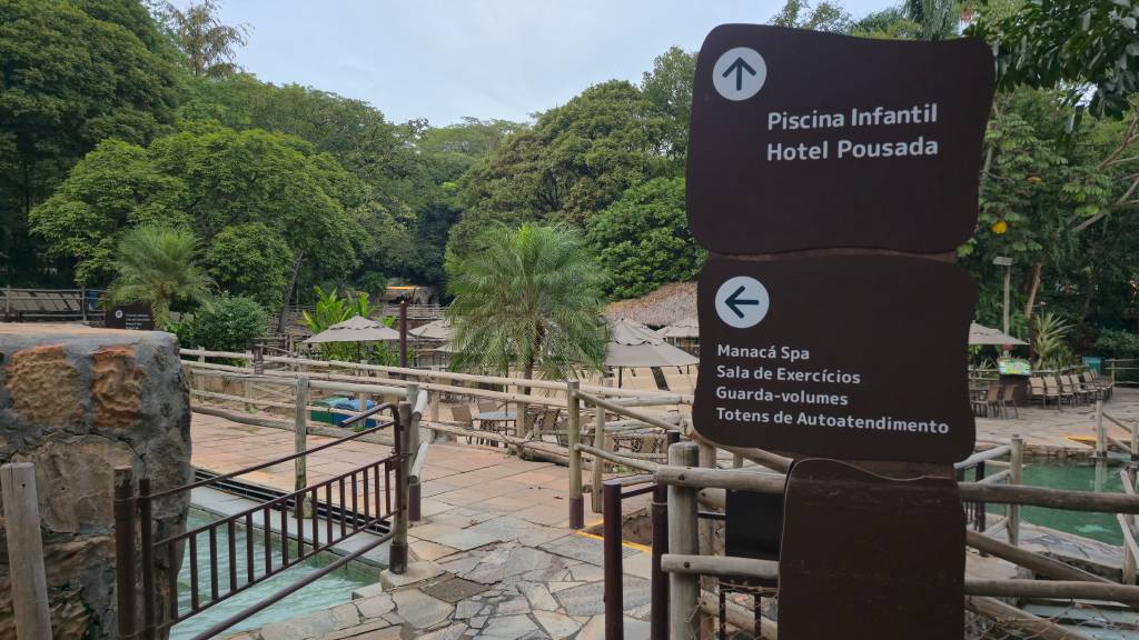 Placa marrom com setas brancas indicando Piscina Infantil Hotel Pousada para cima e Manacá Spa Sala de Exercícios Guarda-volumes Totens de Autoatendimento para a esquerda, em um ambiente externo com árvores, palmeiras, guarda-sóis e piscinas