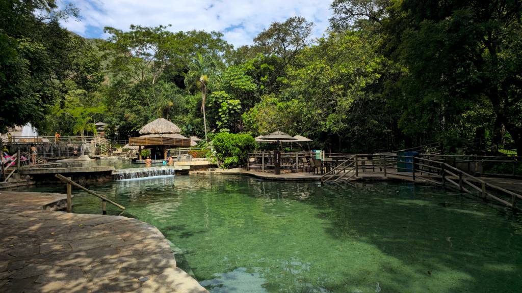 Pessoas relaxam em piscinas naturais de águas cristalinas e esverdeadas, cercadas por densa vegetação tropical. Há passarelas de madeira, quiosques com telhado de palha e mesas com guarda-sóis, sob um céu azul com nuvens esparsas