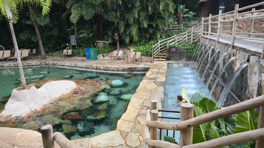 Piscina de águas claras com pedras no fundo e uma pessoa na área de hidromassagem, cercada por vegetação exuberante e uma passarela de madeira com jatos dágua