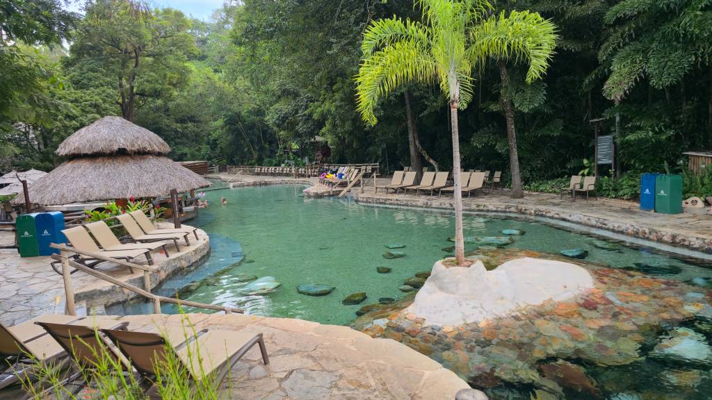 Piscina de águas claras com pedras no fundo, cercada por espreguiçadeiras e vegetação exuberante. Um quiosque com telhado de palha e pessoas na água completam o cenário de lazer.