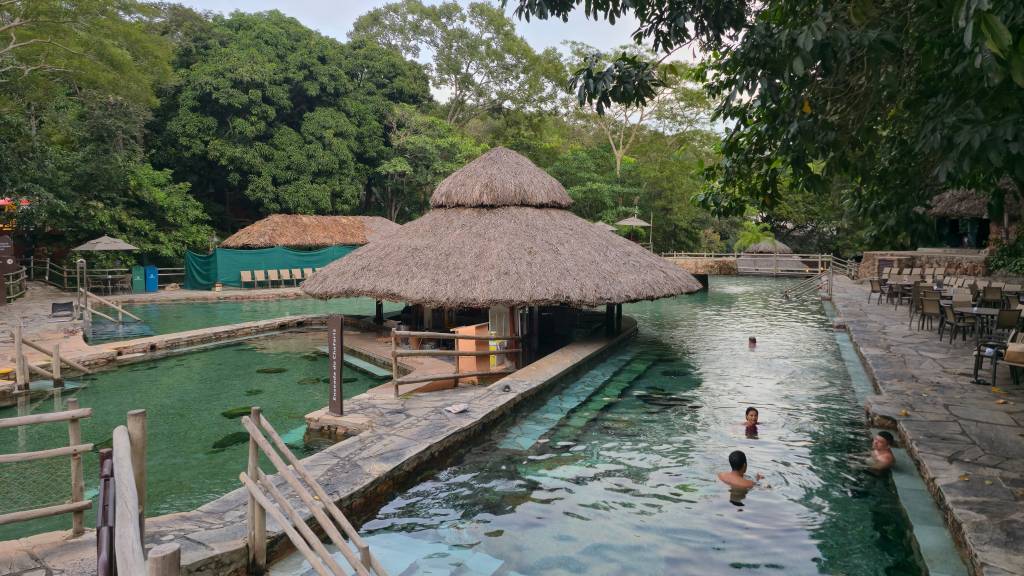Piscina natural de águas cristalinas com pessoas nadando, cercada por vegetação densa e quiosques de palha. Há um bar flutuante no centro e mesas com cadeiras na margem direita, sob a sombra de árvores. O ambiente é de lazer e natureza