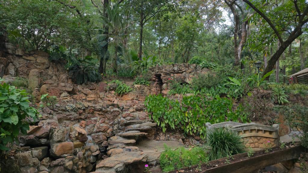 Jardim exuberante com uma cascata de pedras e vegetação densa, incluindo árvores altas e folhagens verdes. Há uma escadaria rústica de pedra e um pequeno túnel na rocha, com um banco de pedra ornamentado à direita.