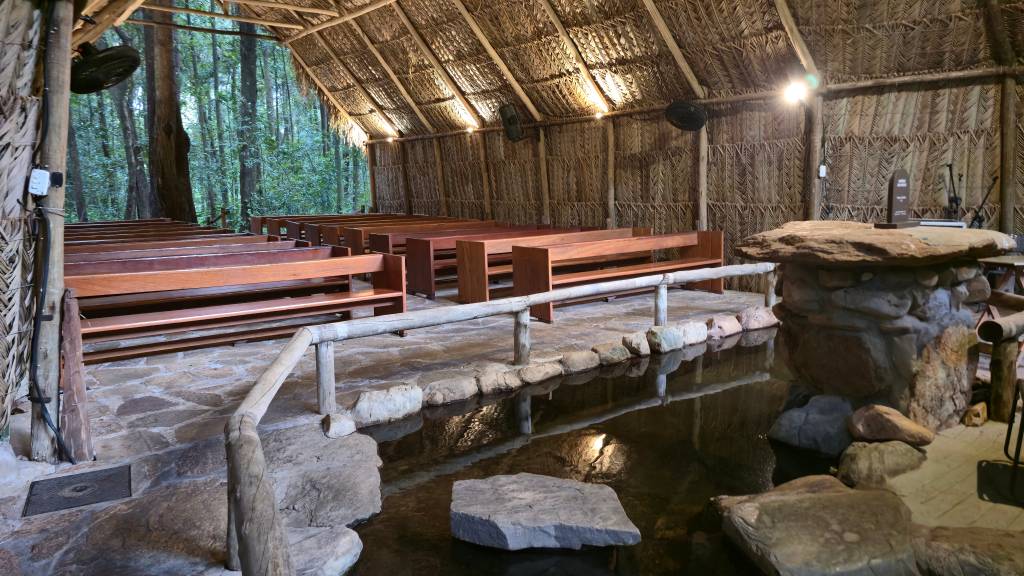 Interior de um templo rústico com teto e paredes de palha, bancos de madeira e piso de pedra. Um espelho dágua com pedras no centro separa os bancos de um altar de pedra. Ao fundo, árvores da floresta.
