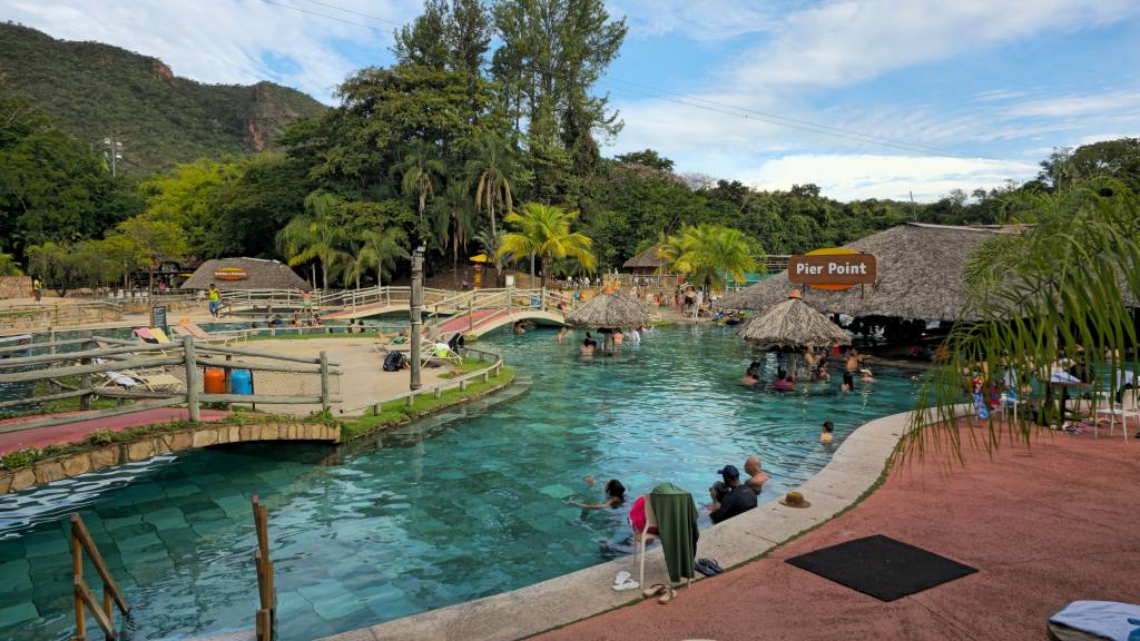 Pessoas nadando em piscinas de águas claras e azuis, cercadas por pontes de madeira e quiosques com telhados de palha. Ao fundo, montanhas verdes e céu azul com nuvens esparsas. Um letreiro indica Pier Point sobre um dos quiosques. O ambiente é de lazer e natureza