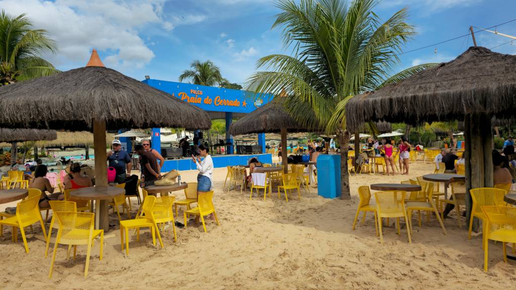 Pessoas relaxam em mesas e cadeiras amarelas sob guarda-sóis de palha em uma praia artificial de areia clara. Ao fundo, um palco azul com a inscrição Praia do Cerrado e palmeiras.