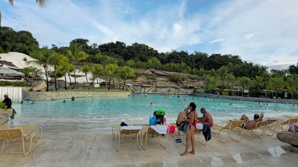 Pessoas relaxam em um parque aquático com piscina de ondas, cercada por coqueiros e vegetação exuberante. Algumas pessoas nadam na água azul-clara, enquanto outras descansam em espreguiçadeiras na área de areia artificial. O céu está parcialmente nublado, e o ambiente é de lazer e diversão