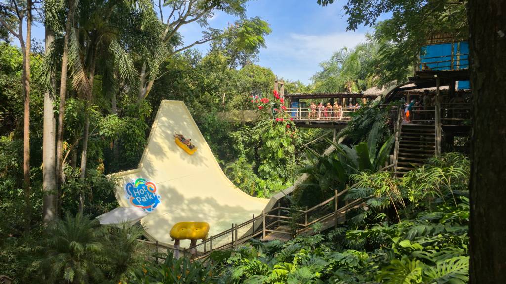 Pessoas descendo um toboágua amarelo em um parque aquático tropical, com o logo Hot Park visível. Ao fundo, uma passarela de madeira com visitantes e vegetação exuberante.