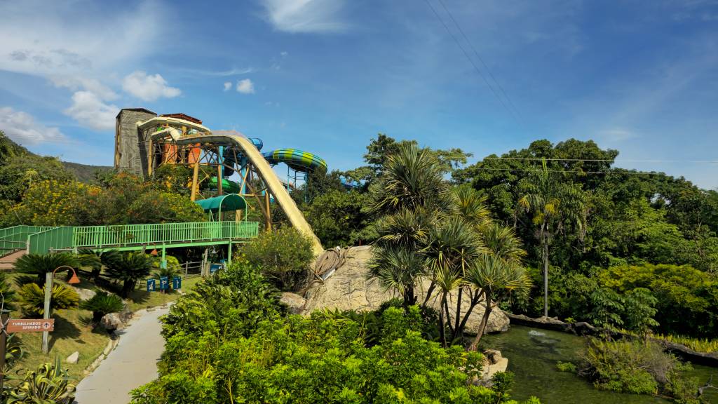 Parque aquático com toboáguas coloridos e uma torre de madeira, cercado por vegetação exuberante e um rio artificial, sob céu azul com nuvens esparsas