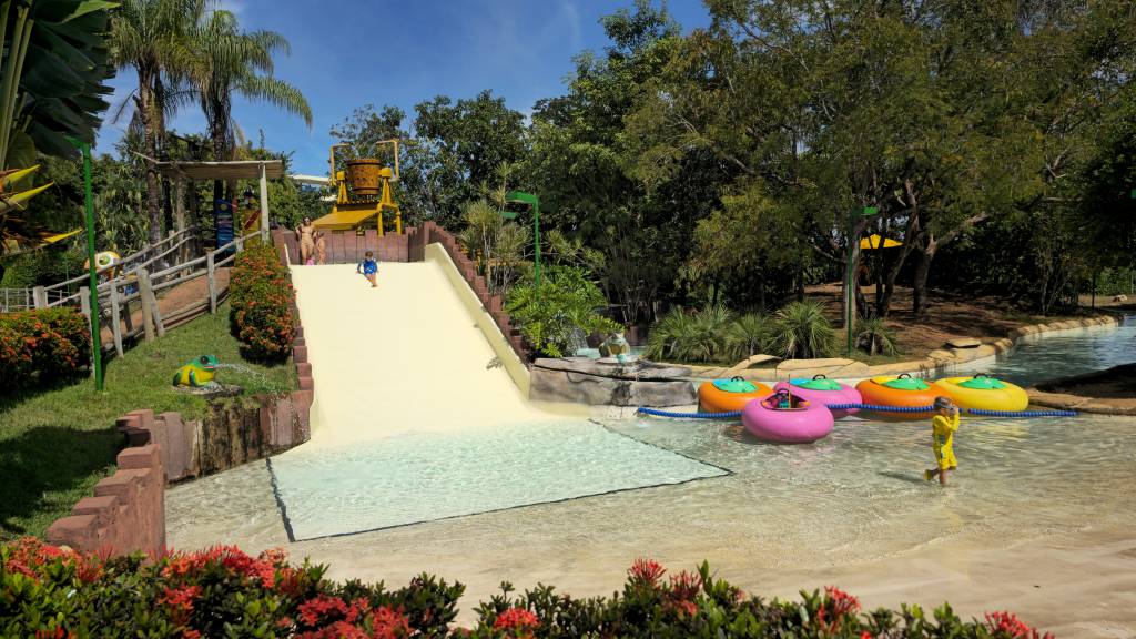 Parque aquático com escorregador bege claro, onde uma criança de camiseta azul desce. Na base, uma piscina rasa com boias coloridas e uma criança de roupa amarela. Ao fundo, árvores e um céu azul claro