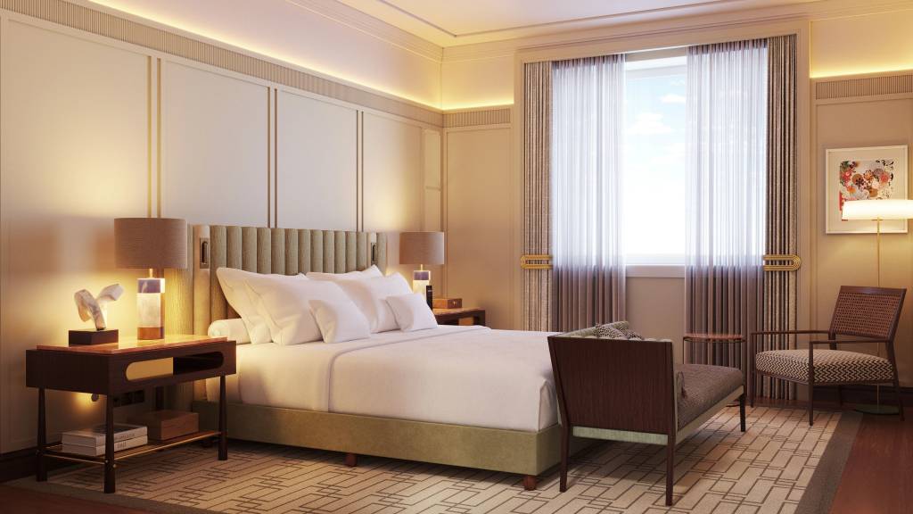 Quarto de hotel luxuoso com cama queen size de lençóis brancos, cabeceira estofada bege, duas mesas de cabeceira escuras com abajures acesos. À direita, uma poltrona de madeira e palha, um banco estofado e uma janela com cortinas claras e escuras. O ambiente tem iluminação quente e um tapete geométrico bege e marrom