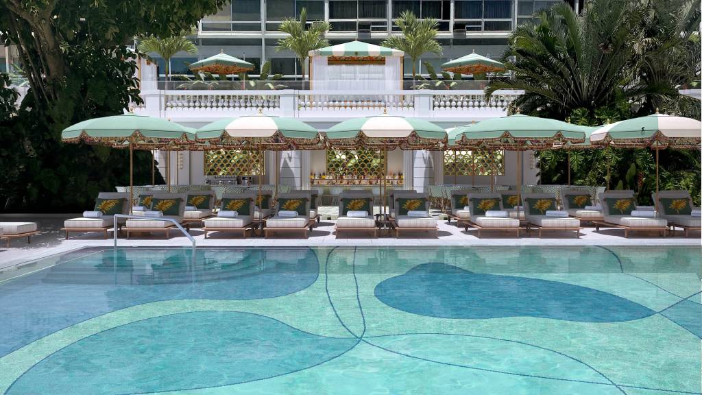 Piscina de hotel luxuoso com azulejos em tons de azul e verde, ladeada por espreguiçadeiras claras com almofadas de estampa tropical e guarda-sóis verde-água. Ao fundo, um edifício branco com varandas e vegetação exuberante