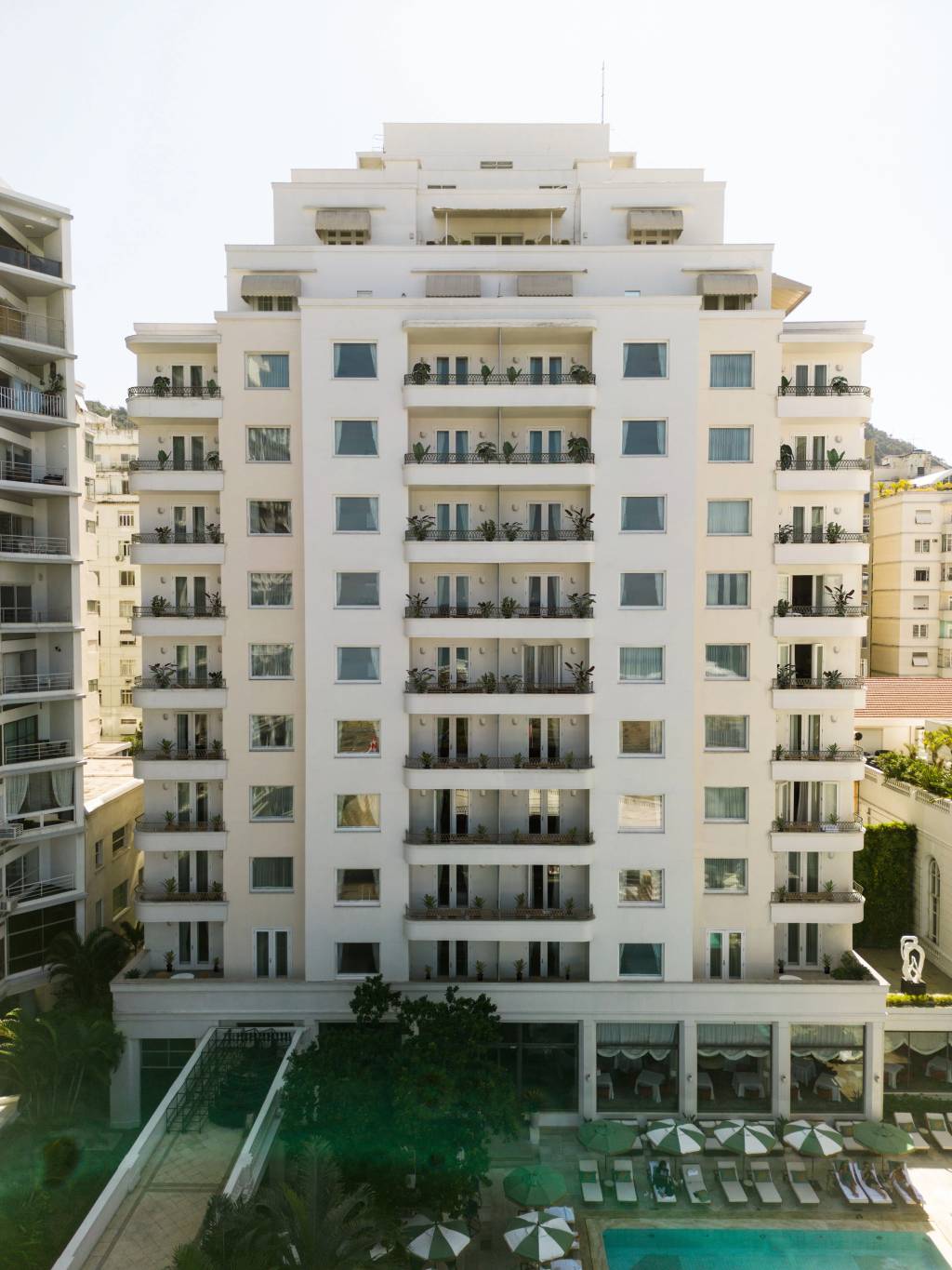 Edifício branco de vários andares com varandas e janelas, algumas com plantas, e toldos bege no topo. Na base, uma piscina com espreguiçadeiras e guarda-sóis verdes e brancos, cercada por árvores e outros prédios ao fundo