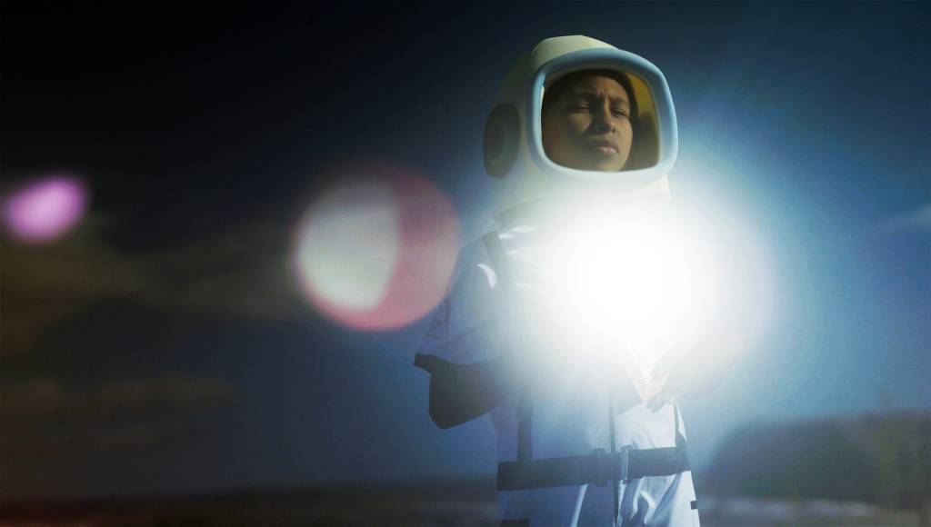 Pessoa de pele morena usando um capacete de astronauta branco e azul claro, com o rosto sério e olhos fechados, em um ambiente escuro com um forte brilho branco à sua frente e pontos de luz coloridos ao fundo