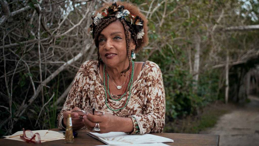 Mulher negra de meia-idade, com turbante de penas e tranças, sorri sentada à mesa de madeira. Veste blusa estampada marrom e branca, colares coloridos e brincos turquesa. Segura óculos na mão, com papéis e um frasco sobre a mesa. Ao fundo, vegetação densa e galhos secos