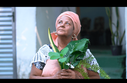 Mulher de pele morena, turbante e blusa salmão, com brincos de argola dourados, segura um buquê de folhas verdes e flores brancas, olhando para cima com expressão pensativa. Ela está em um ambiente externo, com uma parede branca e uma porta de metal ao fundo