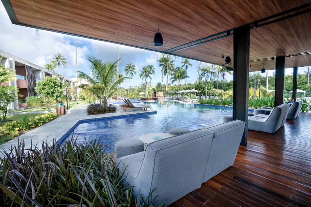 Área de lazer de um resort com piscina azul, palmeiras e espreguiçadeiras sob céu azul com nuvens. À direita, sofás cinzas e mesas redondas em deck de madeira sob teto de madeira. Ao fundo, prédios e vegetação exuberante