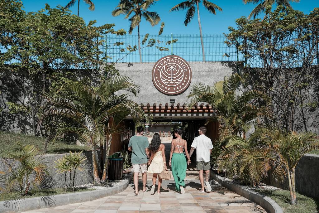 Quatro pessoas, de costas, caminhando em direção à entrada de um resort. A fachada tem um letreiro redondo com Japaratinga Lounge Resort e um coqueiro estilizado. O caminho é de pedras claras, ladeado por grama e palmeiras.