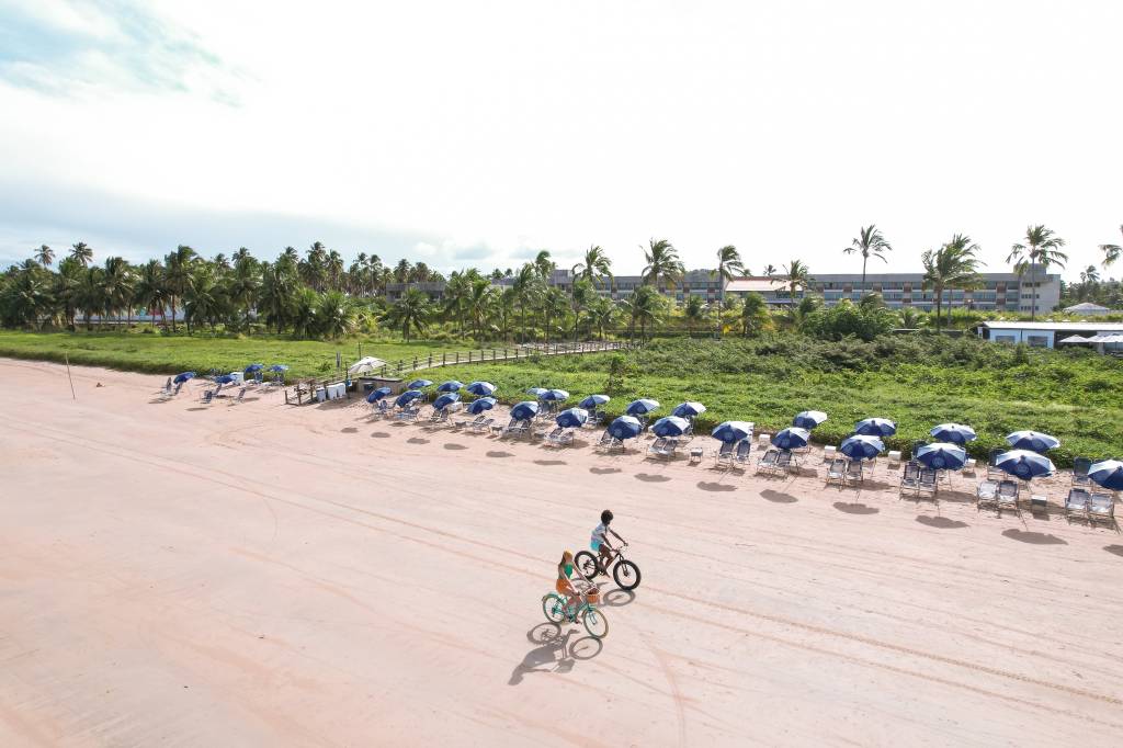 Duas pessoas, uma mulher de blusa laranja e um homem de blusa azul, pedalando bicicletas na areia clara de uma praia. Ao fundo, coqueiros, vegetação densa e um hotel, com fileiras de guarda-sóis azuis e cadeiras de praia na areia. O céu está parcialmente nublado