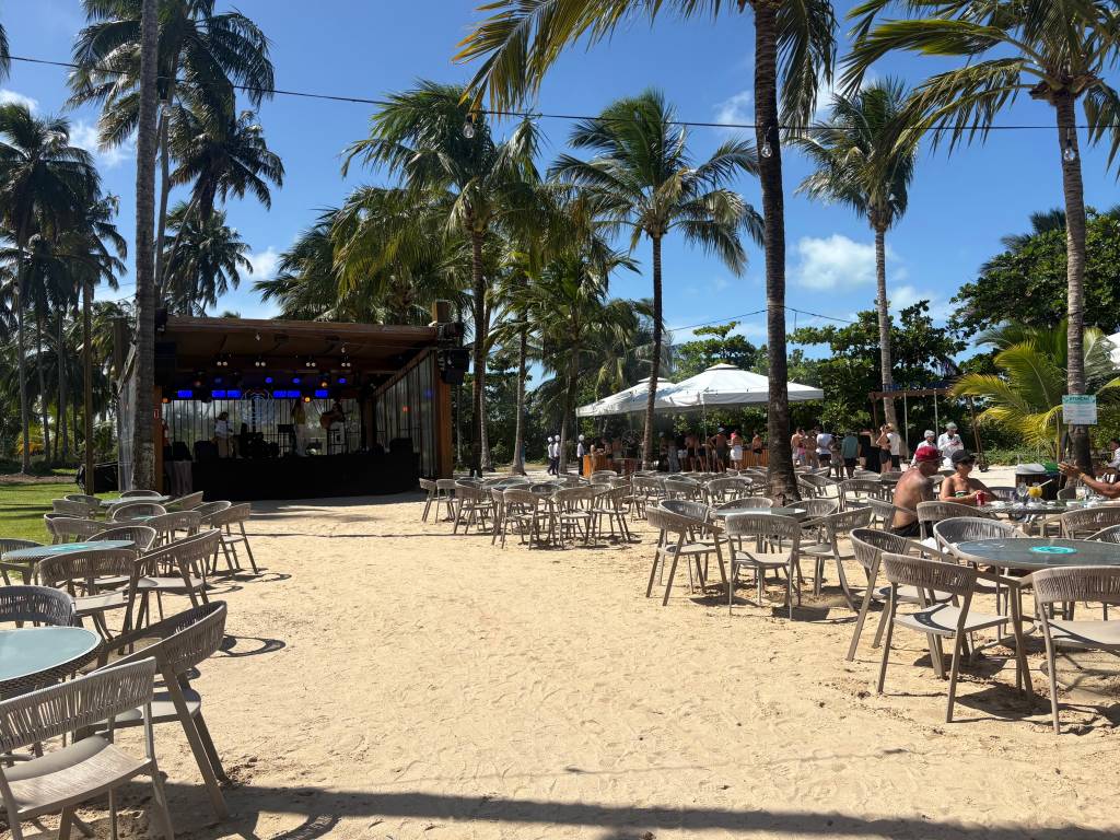 Um palco com iluminação azul e uma banda se apresentando em um bar de praia com mesas e cadeiras de vime na areia, sob coqueiros e céu azul. Pessoas sentadas e em pé, algumas sob um toldo branco ao fundo, desfrutando do dia ensolarado