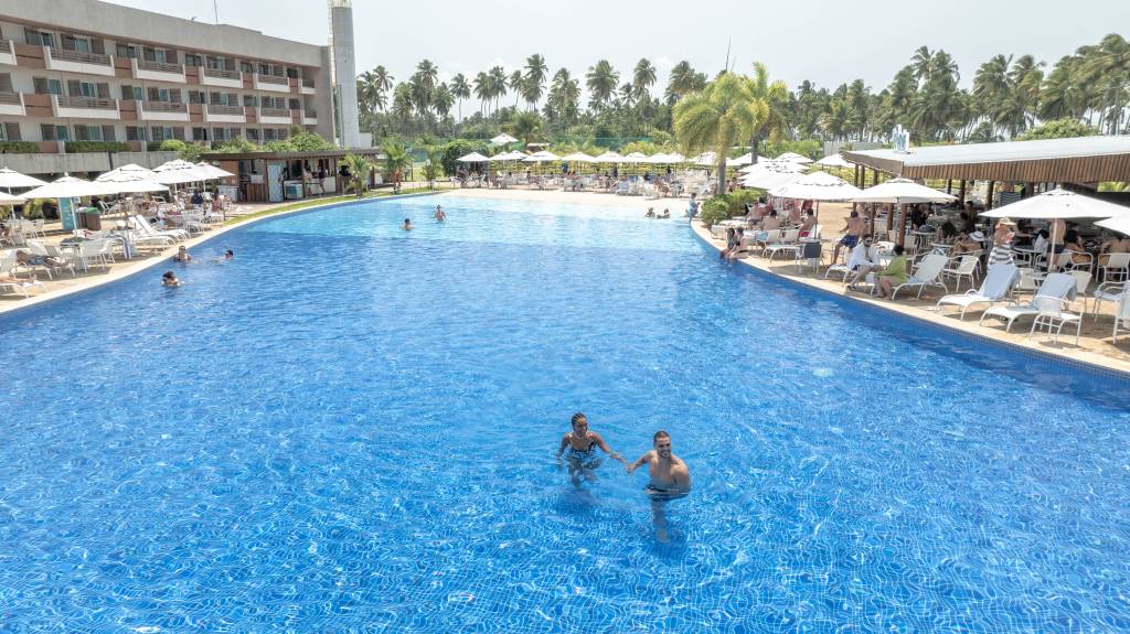 Piscina azul de resort com pessoas nadando e relaxando em espreguiçadeiras sob guarda-sóis brancos, com um prédio de hotel e coqueiros ao fundo, em dia ensolarado