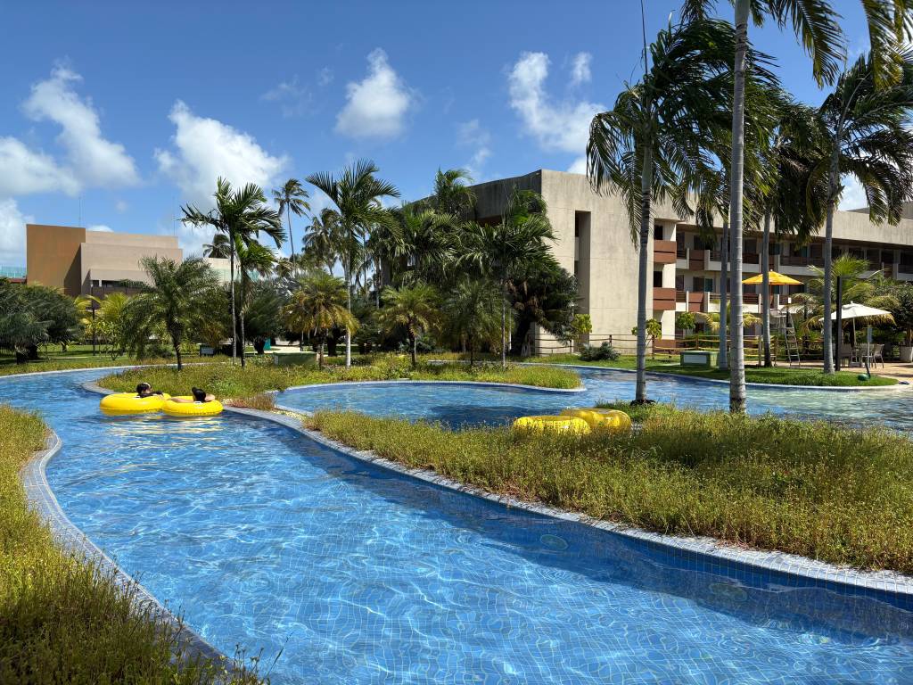 Duas pessoas relaxam em boias amarelas em um rio lento de piscina azul, cercado por grama e palmeiras. Ao fundo, prédios de hotel e céu azul com nuvens brancas, sugerindo um resort tropical ensolarado