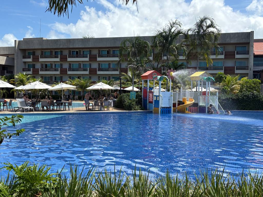 Piscina de resort com água azul, escorregador infantil e jatos dágua. Ao fundo, um prédio de vários andares, palmeiras e pessoas relaxando sob guarda-sóis brancos em mesas e cadeiras, sob céu azul com nuvens