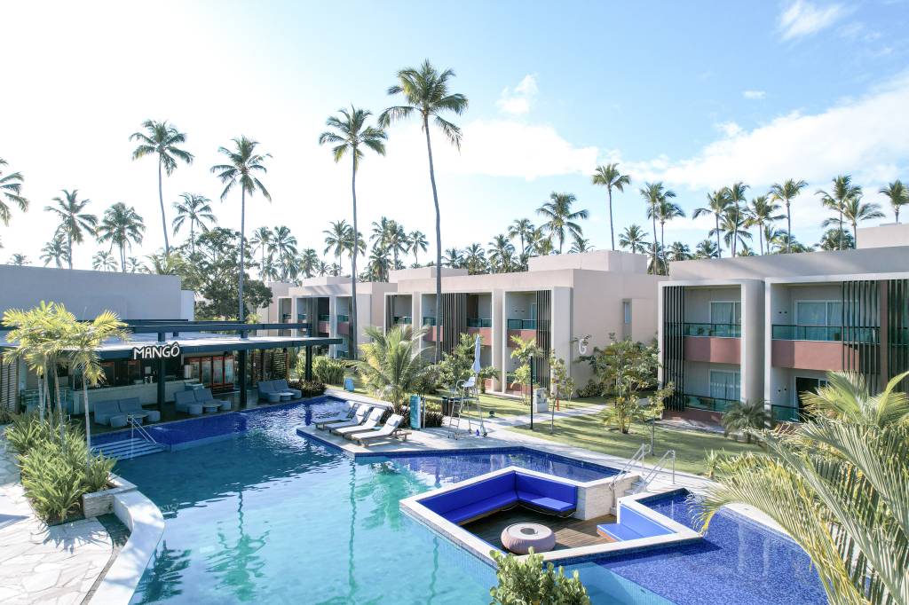 Resort tropical com piscina azul, espreguiçadeiras e um bar chamado Mango à esquerda. Ao fundo, prédios de apartamentos bege com varandas e muitas palmeiras altas sob um céu azul claro com nuvens esparsas