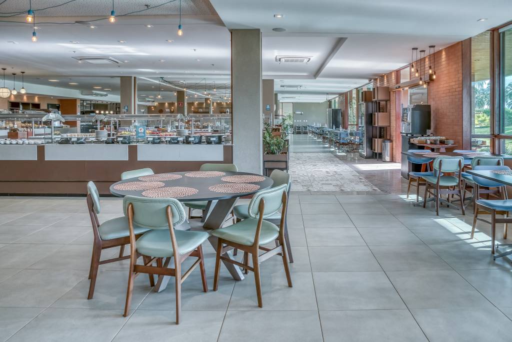Salão de restaurante amplo e moderno, com mesas redondas e cadeiras estofadas em tons de verde e azul. Ao fundo, um buffet com diversas opções de alimentos e uma área de refeições com janelas grandes que revelam a vegetação externa