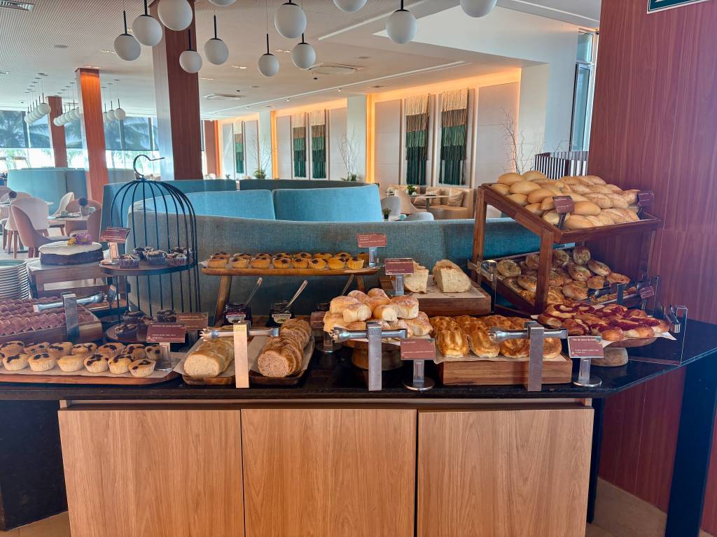 Mesa de buffet com pães variados, bolos, tortas e doces em um salão iluminado, com sofás azuis e mesas ao fundo