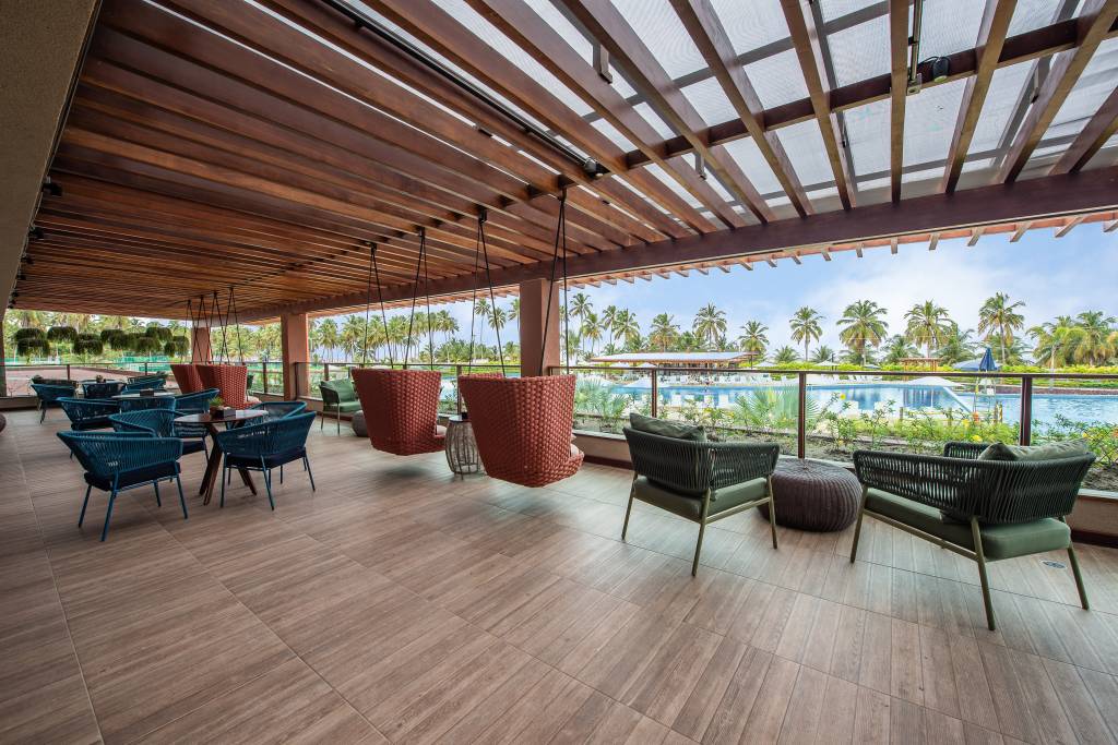 Varanda de um resort com teto de madeira e piso claro, mobiliada com cadeiras e mesas azuis, poltronas suspensas de vime laranja e poltronas verdes com almofadas. Ao fundo, uma piscina azul e coqueiros sob céu claro