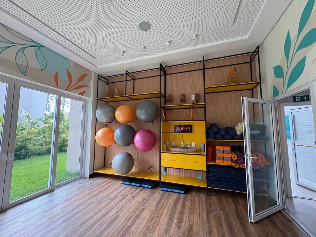 Sala de ginástica com piso de madeira, parede decorada com folhas e prateleiras amarelas. Há bolas de pilates coloridas, tapetes de yoga e um relógio digital exibindo 09:08:23. Uma porta de vidro aberta revela um jardim gramado