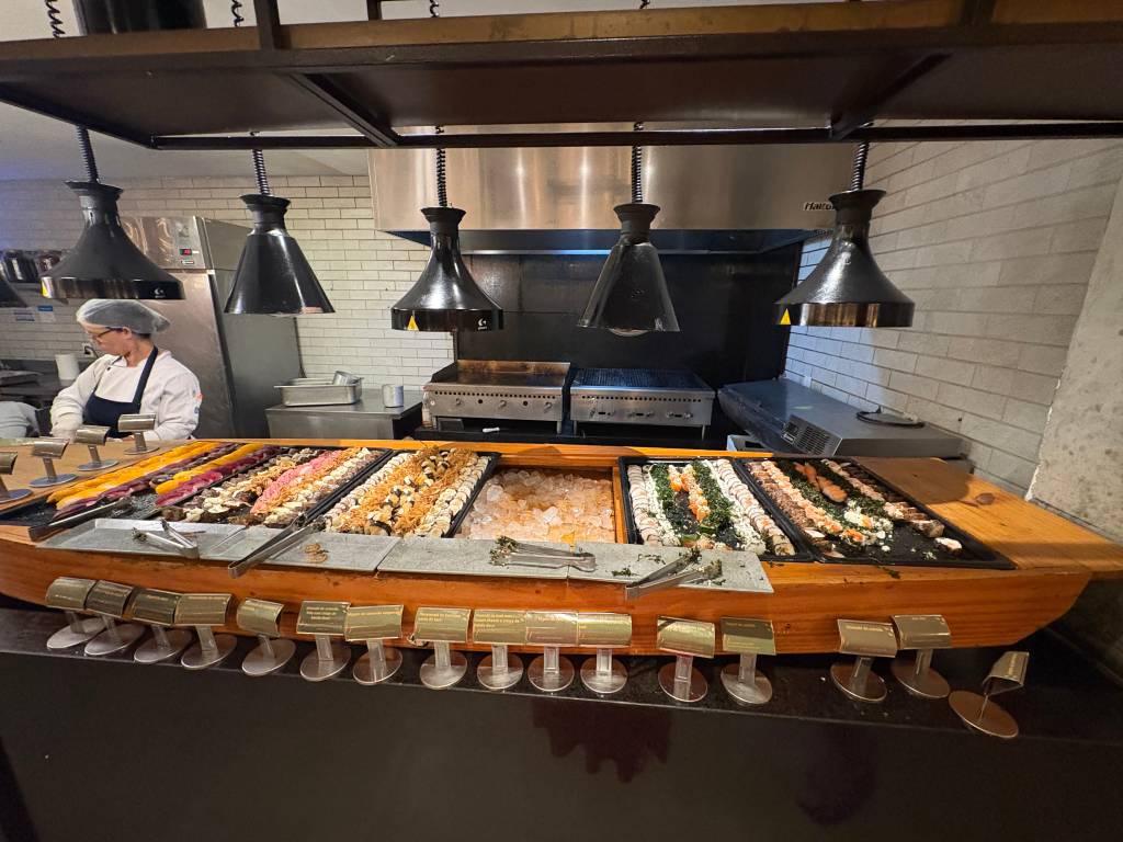 Buffet de sushi com uma variedade de makis coloridos e niguiris em bandejas pretas sobre gelo, dispostos em um balcão de madeira em formato de barco. Uma chef de touca e uniforme branco e avental azul trabalha ao fundo, sob lâmpadas pretas penduradas.
