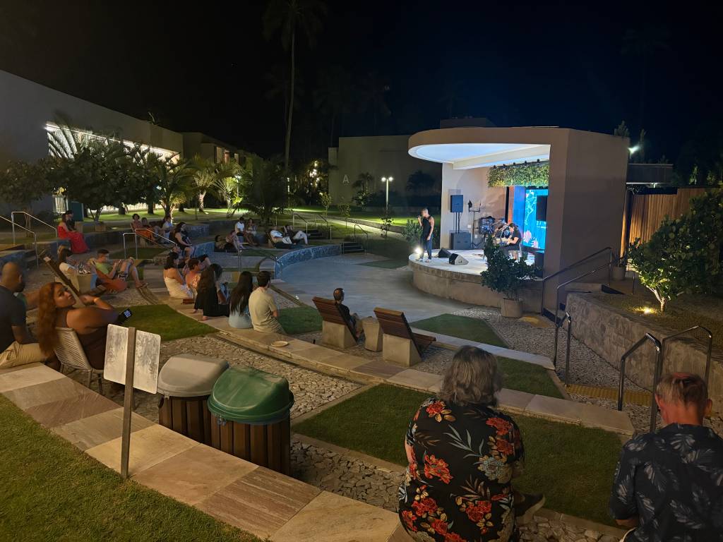 Pessoas assistem a um show noturno em um anfiteatro ao ar livre, com um cantor no palco iluminado e um telão azul ao fundo. A plateia está sentada em degraus de pedra e cadeiras de madeira, cercada por gramados e palmeiras sob o céu escuro