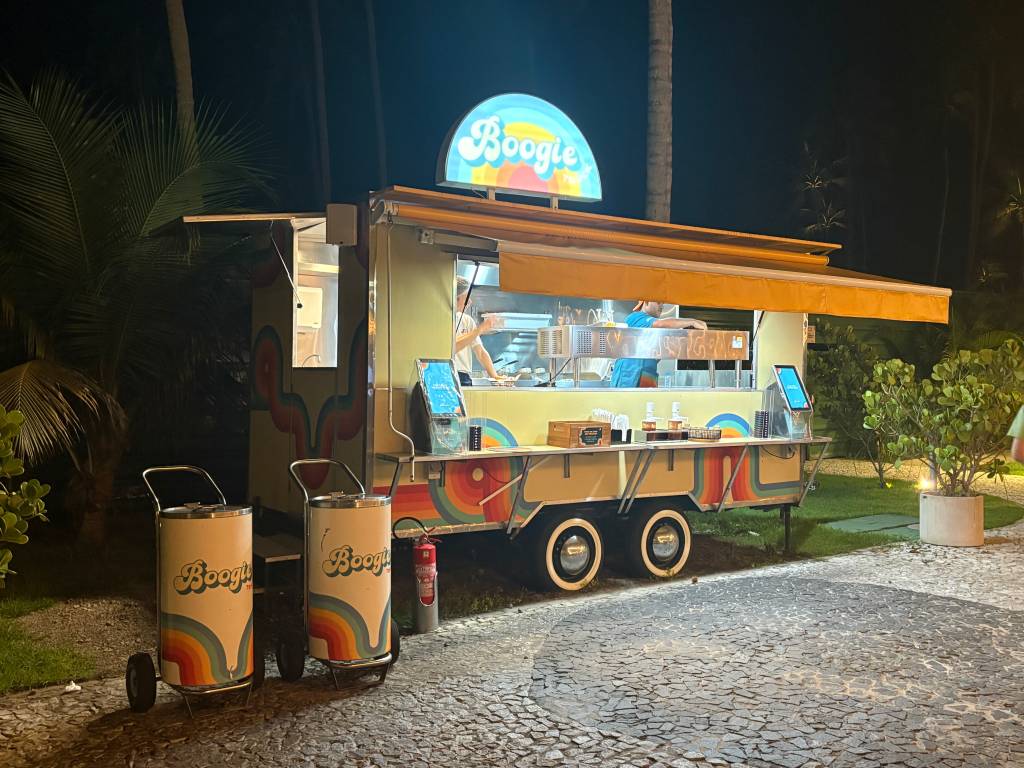 Food truck amarelo e bege com toldo laranja e o nome Boogie em azul claro, iluminado à noite. Um funcionário de camisa azul prepara alimentos dentro do veículo, que tem detalhes de arco-íris e está estacionado em um piso de pedras portuguesas, com vegetação ao redor. Dois carrinhos de lixo com o mesmo design estão à frente do food truck