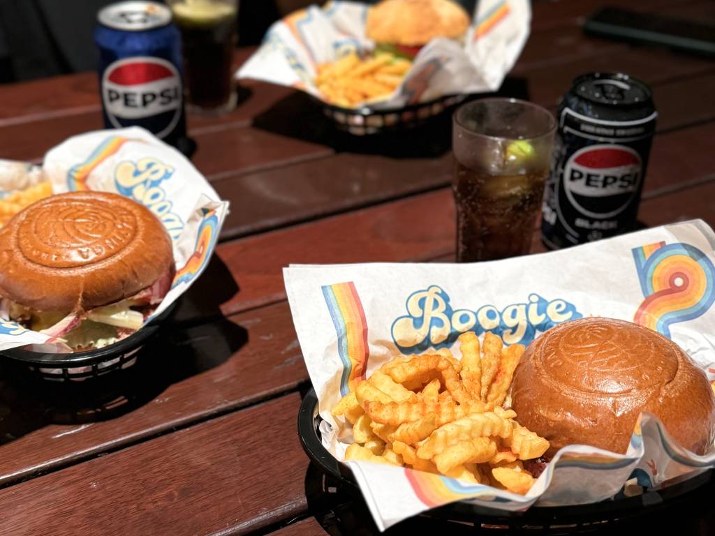 Três combos de hambúrguer com batatas fritas e latas de Pepsi em uma mesa de madeira escura. Dois combos estão em primeiro plano, um com Pepsi Black e outro com Pepsi tradicional. O terceiro combo está desfocado ao fundo