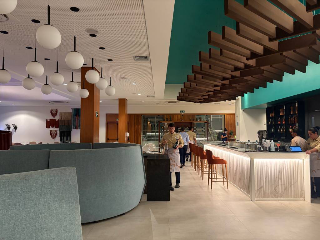 Interior de um restaurante moderno com teto branco pontilhado e luminárias esféricas brancas penduradas. À direita, um balcão de bar iluminado com banquetas marrons e funcionários atendendo. No centro, um garçom de camisa bege e avental branco caminha carregando garrafas. Sofás curvos azuis e mesas de jantar ao fundo completam o ambiente.