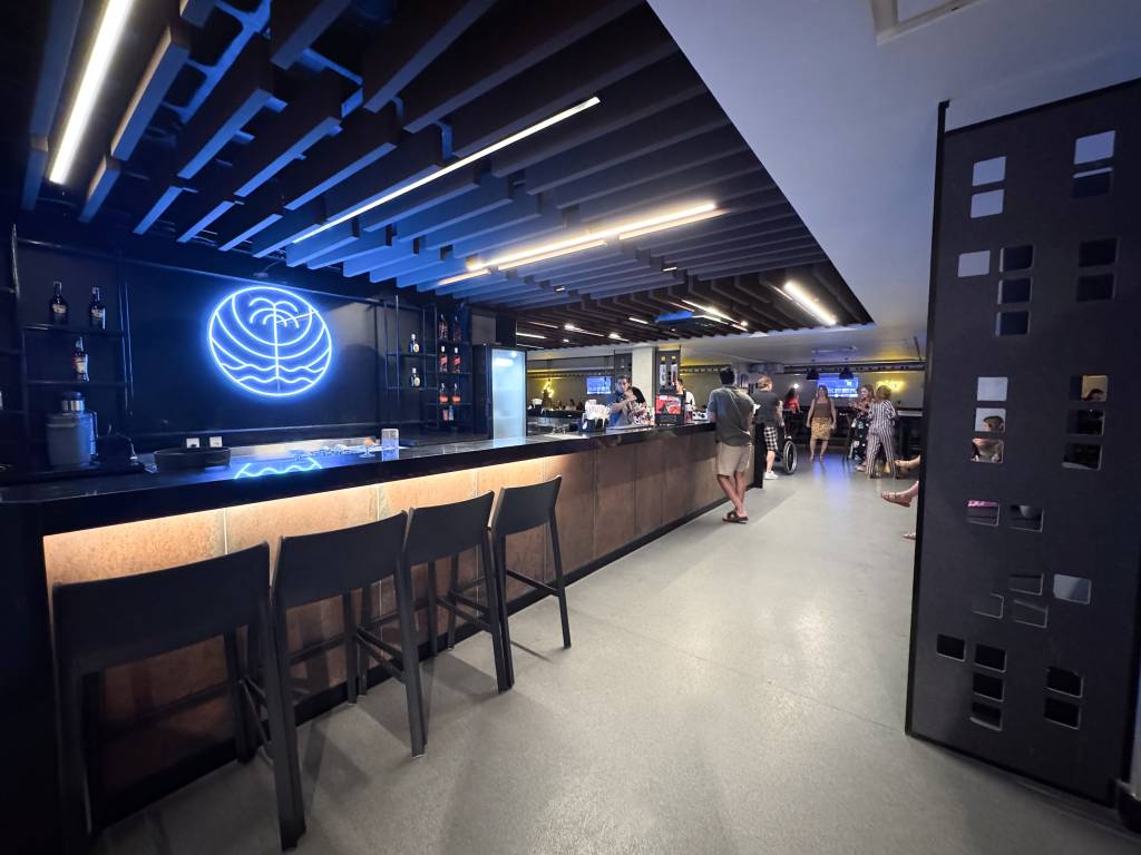Interior de um bar moderno com balcão longo e quatro banquetas pretas. Ao fundo, um letreiro neon azul com um coqueiro estilizado e pessoas conversando e circulando no ambiente iluminado