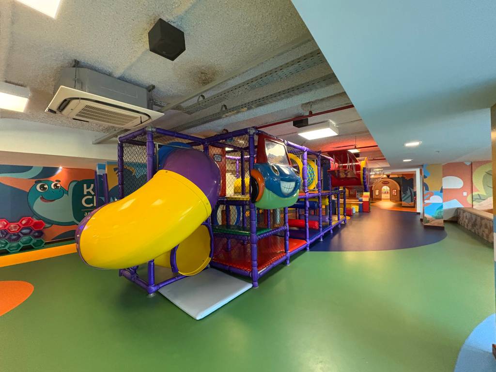 Playground infantil colorido com escorregador amarelo e roxo, estrutura de escalada e um carro azul de brinquedo. O chão é verde e azul, e as paredes têm desenhos de personagens infantis. Ao fundo, um túnel e uma casinha de madeira.